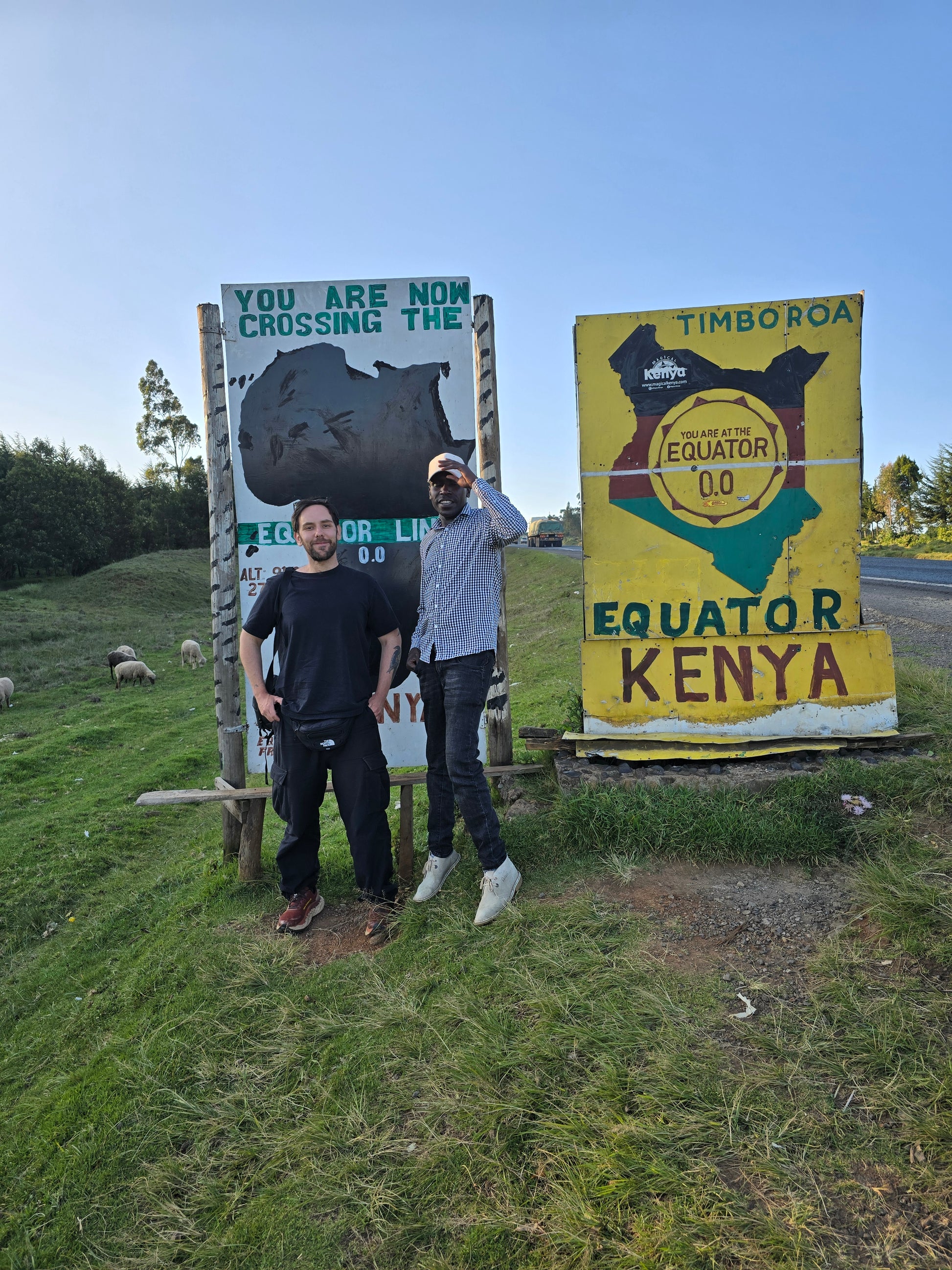 Two people standing in front of a large sign with text and graphics on a grassy area.