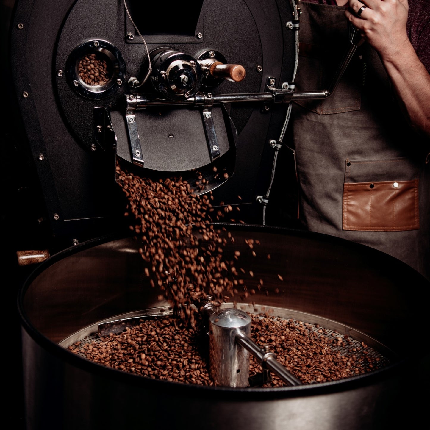 Coffee beans being poured from a coffee roaster into the roasters cooling tray. A person, the roaster, is standing by the roaster wearing a denim apron