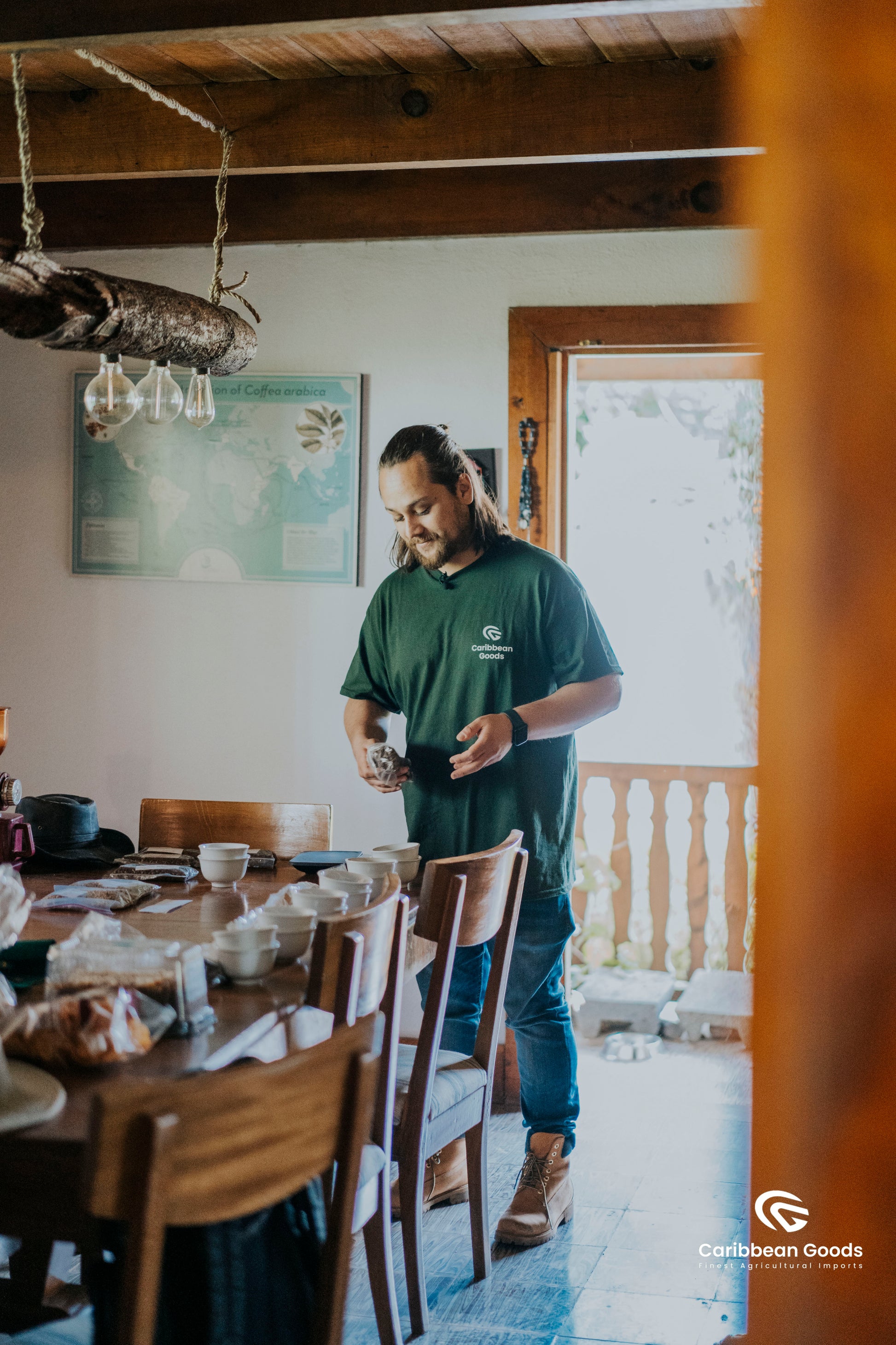 Man in a green shirt standing in a dining room setting up for a coffee cupping session, with wooden furniture and a map on the wall.