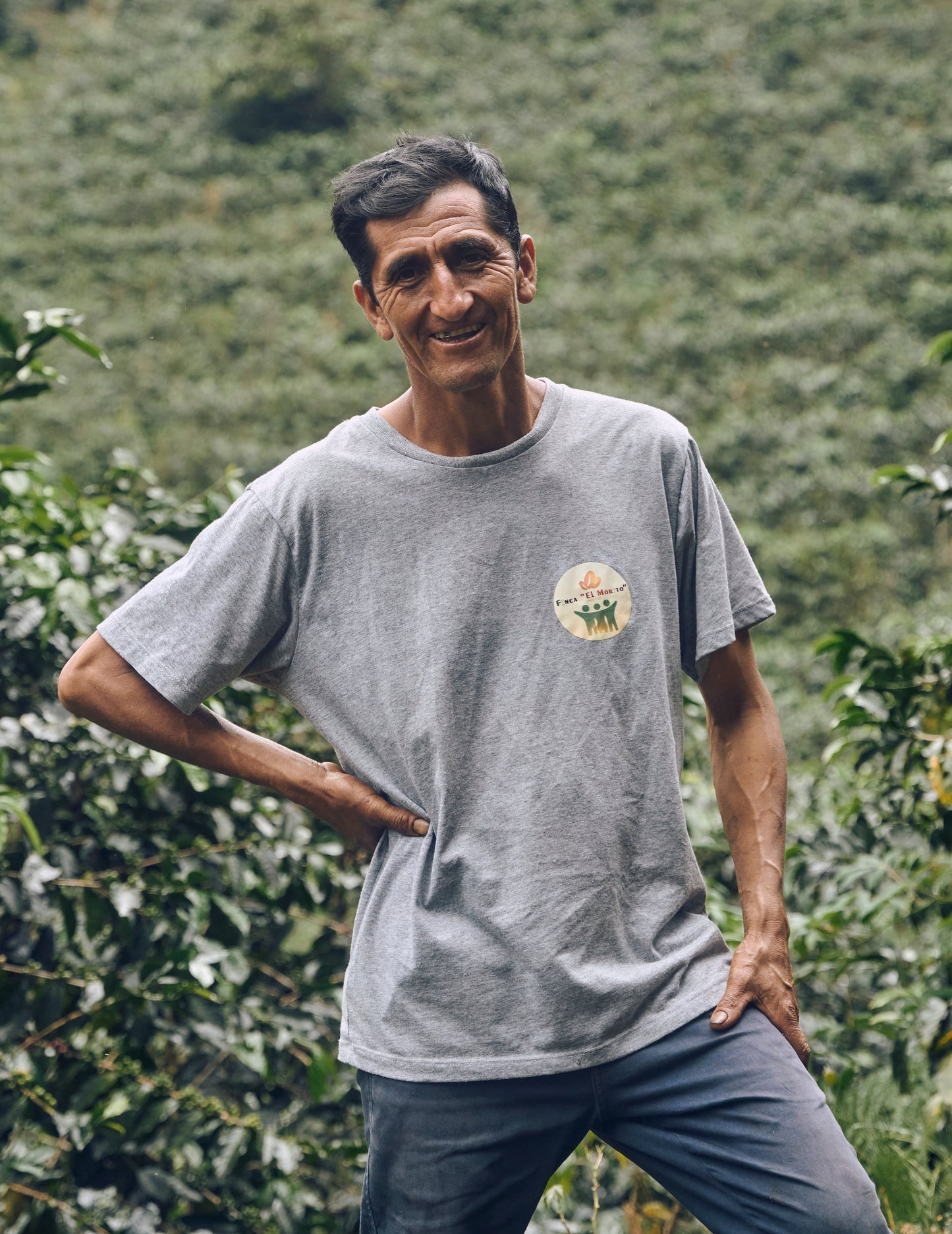 Man standing in a coffee plantation wearing a gray t-shirt with a logo.
