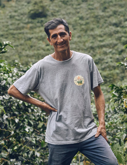 Man standing in a coffee plantation wearing a gray t-shirt with a logo.