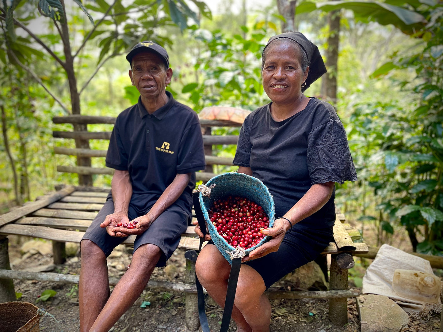 Two people sitting outdoors with a basket of red berries, surrounded by greenery.