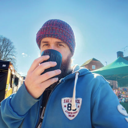 Chris wearing a blue hoodie and colourful knit hat, holding a black huskee cup outdoors on a sunny day.