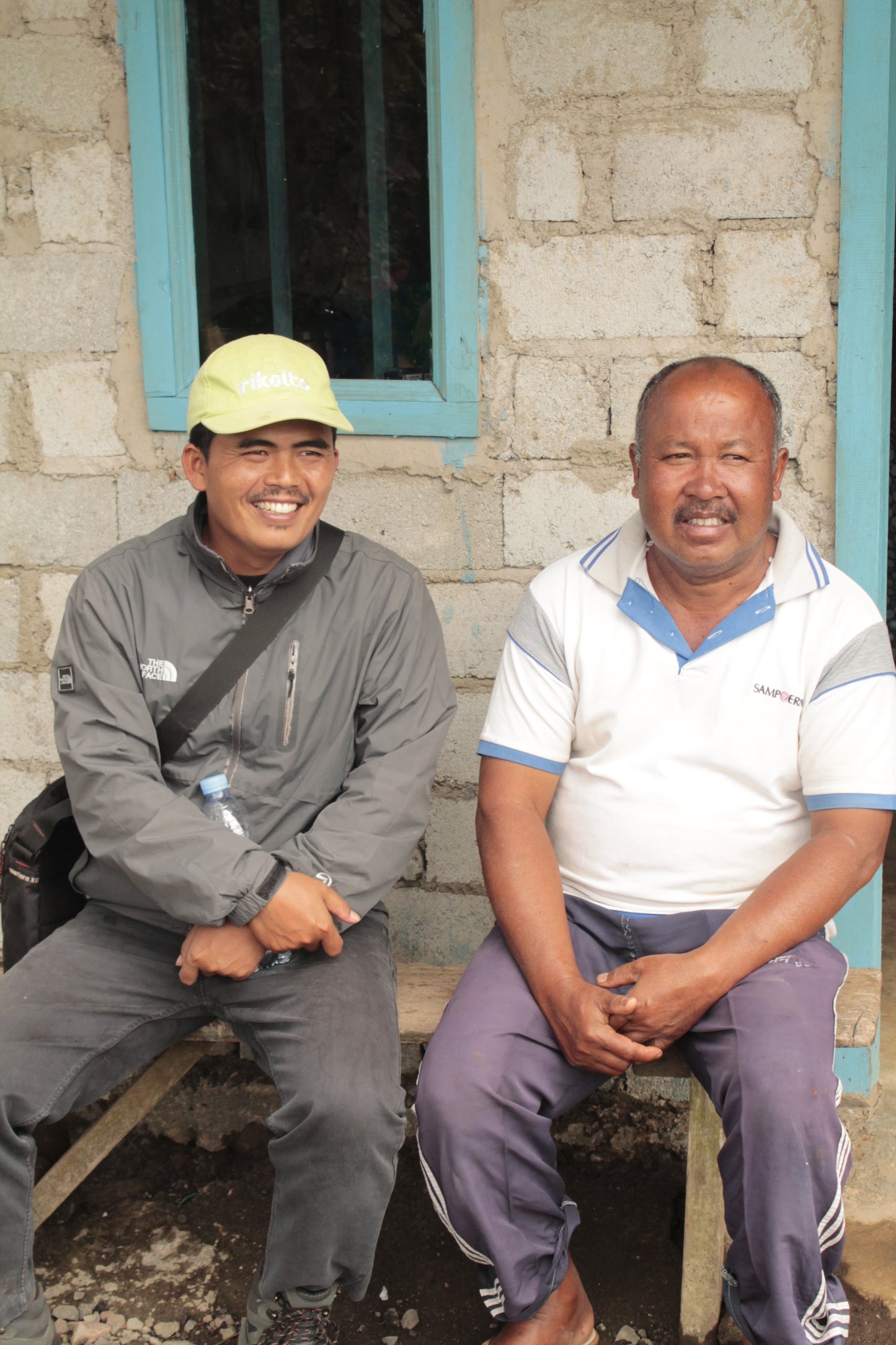 Two men sitting on a bench outside a building with a stone wall.