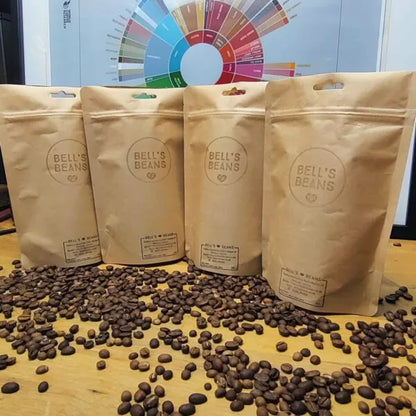 Four bags of 'Bell's Beans' coffee on a wooden surface with scattered coffee beans, and a color wheel in the background.