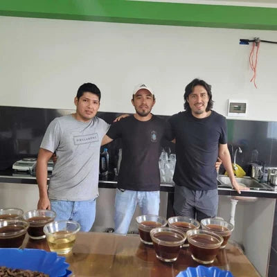 Three men standing together in a kitchen or bar setting with several glass cups of coffee arranged on a wooden table in the foreground.