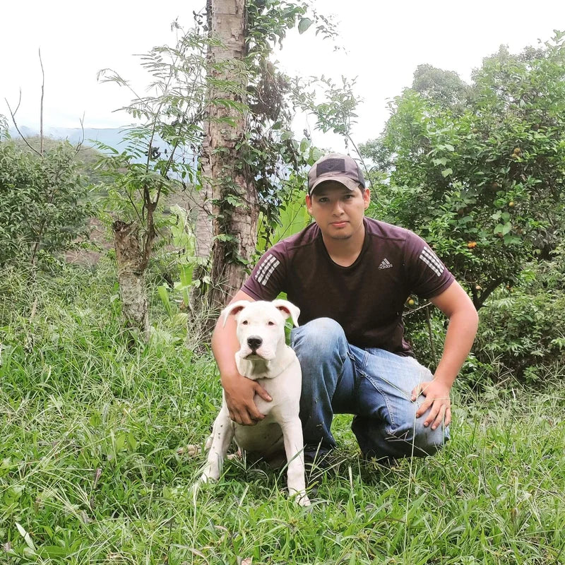 A man, Winker Tapid, wearing a dark brown Adidas t-shirt and blue jeans kneels in a grassy outdoor setting next to a white dog.