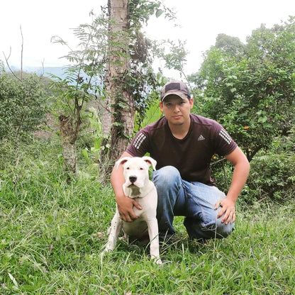A man, Winker Tapid, wearing a dark brown Adidas t-shirt and blue jeans kneels in a grassy outdoor setting next to a white dog.